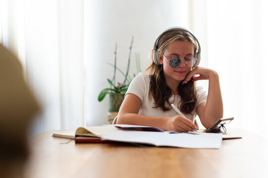 A teenage girl wearing eyeglasses with an eye patch for strabismus treatment does homework indoors while using headphones. She writes in a notebook with a phone placed nearby.