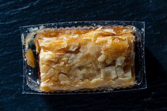 Single piece of Greek baklava in plastic packaging. Top view closeup of sweet golden crispy baklava pastry inside clear plastic container placed on dark stone background