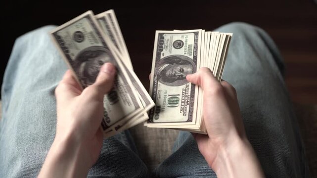 Money. A young man is counting stacks of money, holding multiple bills in his hands. The young man focuses on the money while seated comfortably.