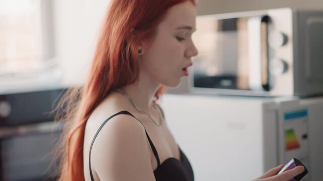 Soft window closeup caucasian woman holding cosmetic bottle, reading label with delicate necklace, thoughtful gaze and natural makeup, intimate morning skincare ritual against warm kitchen backdrop