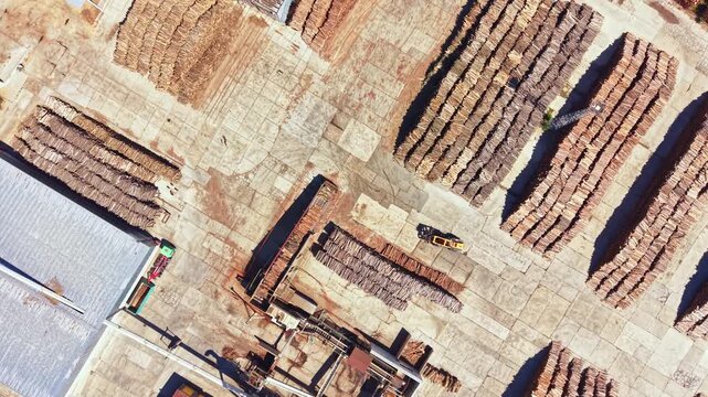 Large area shows stacks of timber arranged in rows. Machinery is visible on site, and the ground has a dirt surface. This location is busy with wood processing activities.