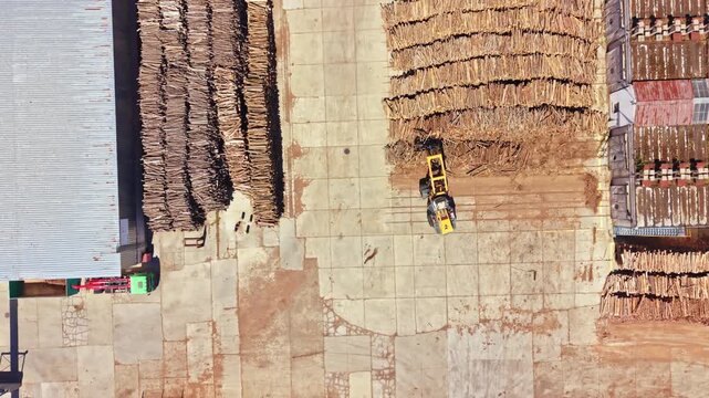 Workers are processing wood at a facility. Large piles of logs are stacked in organized rows. A machine is moving logs around the yard. The scene shows the work being done.