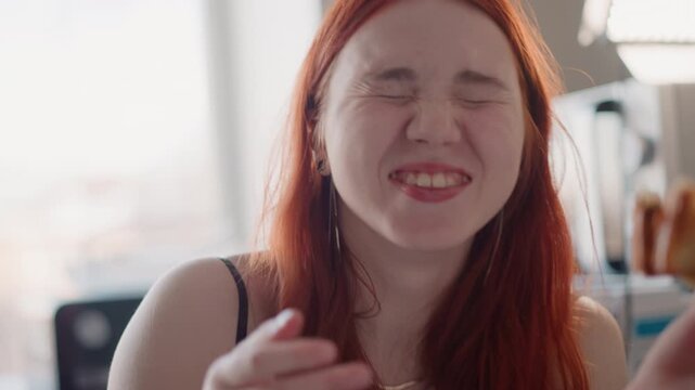 Young woman eating pastry in kitchen, playful morning closeup shows smiling redhead savoring flaky pastry, gentle sunlight, casual outfit, relaxed expression, fingers messy with crumbs, candid