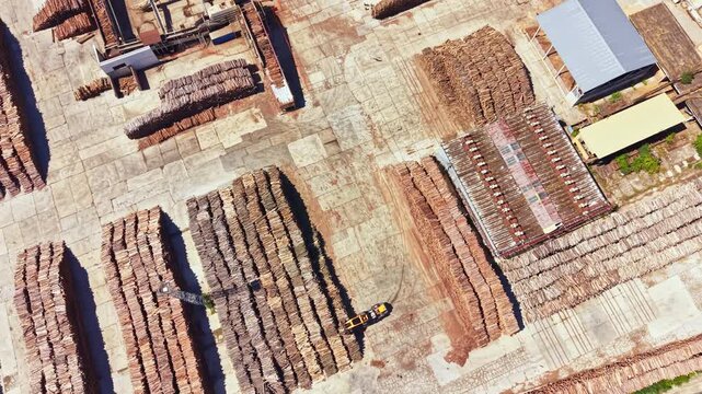 Aerial view of a wood processing area. Stacked logs are organized in rows. A vehicle moves through the site. The location is busy with wood materials ready for use.