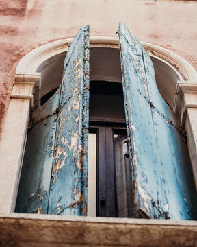 View of an arched window with weathered blue wooden shutters and peeling paint set in a rustic stone wall in Venice, Veneto, Italy.