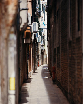 View of a narrow, quiet alleyway with historic brick walls, air conditioning units, and a red fire hydrant in the shadows of a sunny day in Venice, Veneto, Italy.