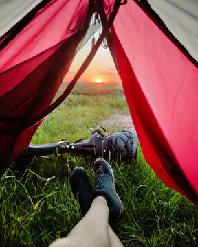 View of a person relaxing inside a tent looking out at a grassy field during a vibrant sunset with a bicycle and gear nearby in Poland.