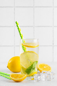 Refreshing homemade lemonade served in clear glass with fresh lemon slices, mint leaves, ice cubes and paper straws on a light kitchen counter against a white tile background with copy space.