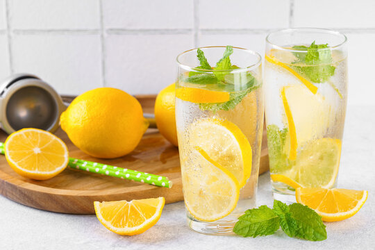 Refreshing homemade lemonade in glasses with fresh lemon slices, mint leaves, ice cubes, sparkling water served on wooden tray on white tiled kitchen background. Summer citrus beverage, drink concept.