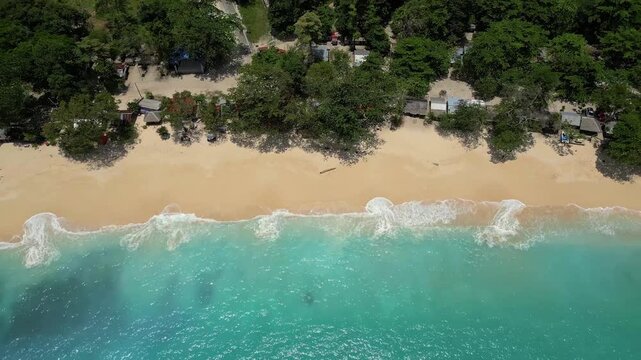 Aerial view of Paal Beach featuring turquoise waves crashing on the sandy shore with lush trees and coastal structures in Likupang Timur, North Sulawesi, Indonesia.
