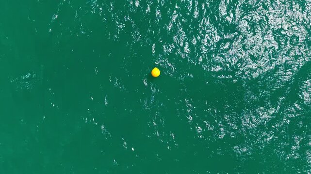 A yellow buoy floats in deep green water. The water reflects sunlight and has subtle waves. This scene is likely near a coastal area or harbor.