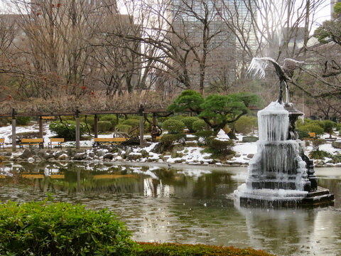 View of a frozen fountain with a crane statue surrounded by snow-covered grounds, bare trees, and park benches in Hibiya Park, Tokyo, Tokyo, Japan.