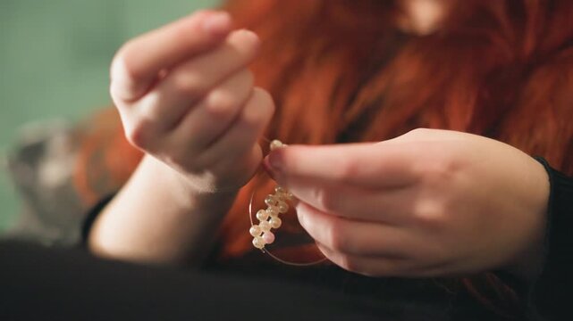 Closeup caucasian fortune teller hands threading beads, red hair blurred background, delicate fingers manipulating crystal bracelet, intimate ritual scene, contemplative atmosphere with warm vintage