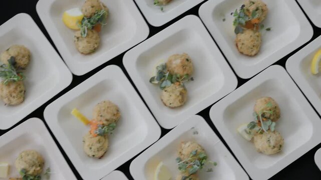 Overhead view of crab cakes topped with microgreens and orange aioli served with lemon wedges on white square plates arranged in rows on a black table at a catered event