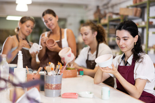 During master class, female participant draws pattern on walls of white ceramic bowl with brush. Girl paints blank, makes individual order. Hand made art, creative pastime.