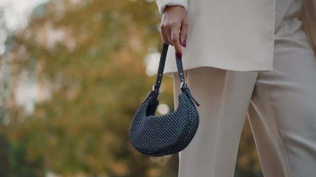 Waist to thigh view of woman walking outdoors, holding small studded handbag by braided strap while bag sways beside tailored blazer and cropped trousers in soft daylight, with handbag, walking