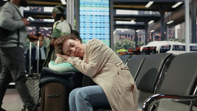 Fatigued woman sleeps on a neck pillow in a bus station during a long wait between routes, dozing on luggage in the waiting area before continuing her journey.