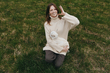 Fototapeta premium Cheerful woman outdoors holding a goat kid in a sunlit field, cozy sweater, relaxed smile, gentle breeze, natural colors, lush grass, rural lifestyle moment