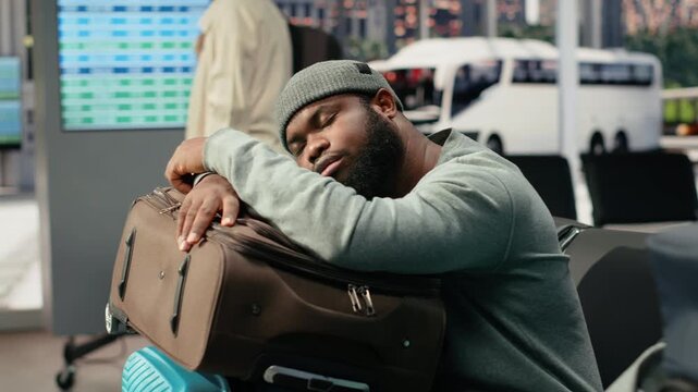 Tired man rests on luggage in a bus station during a long wait for a delayed shuttle, leaning on a travel bag to recover before continuing a difficult trip toward another country after the delay.