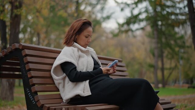 Mature woman in coat and shawl sits on curved wooden bench in city park, lifts smartphone to ear and adjusts hair while crossed legs and calm posture suggest relaxed conversation in soft overcast