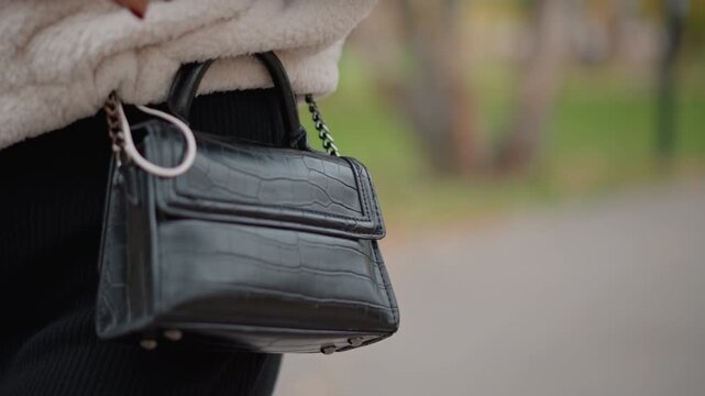Small black croc-embossed handbag swings at hip while person walks along softly blurred park pathway, with glossy textured leather, visible zipper pocket, D-ring hardware, neat stitching, and