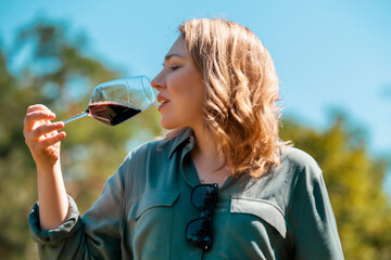 Naklejka premium Woman smelling red wine in glass outdoors on sunny day, in green shirt and sunglasses. Wine tasting experience and relaxing weekend leisure.