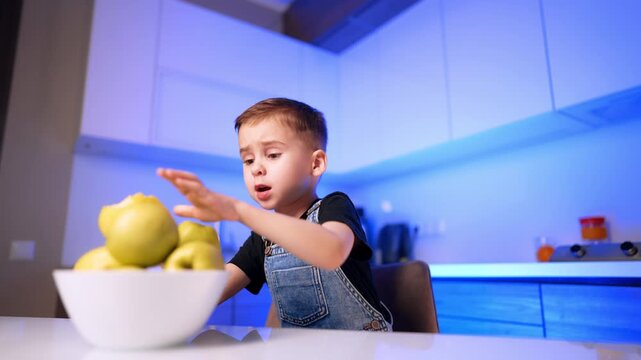 Boy reaching for bowl of apples on table. A child sitting indoors extends his arm toward fresh yellow fruit in a kitchen illuminated with blue light.
