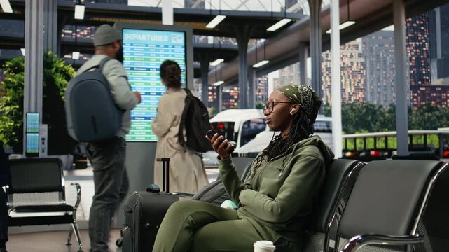Smiling traveler uses a smartphone in a bus terminal to send audio messages, recording voice notes on a mobile phone while sitting on a bench in the coach terminal during the wait.