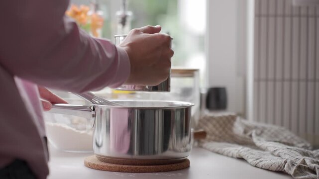 Woman sifts flour into metal saucepan on kitchen counter. Concept of cake batter preparation and cooking ingredients