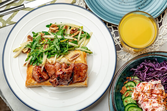 Food served on a plate with green salad. A meal is arranged on a table with a flaky pastry and green salad. There are drinks and side dishes present as well.