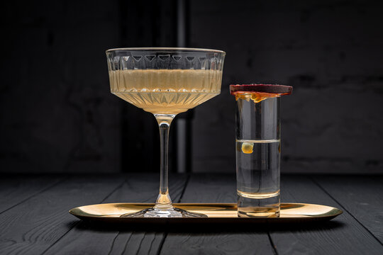 Cocktail coupe and shot glass on metal tray. Yellow cocktail in coupe glass beside shot glass garnished with passionfruit slice on metal tray.