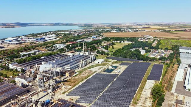 A large industrial complex is near a river. Solar panels cover a nearby field. Green fields and buildings fill the surrounding area under a clear blue sky.