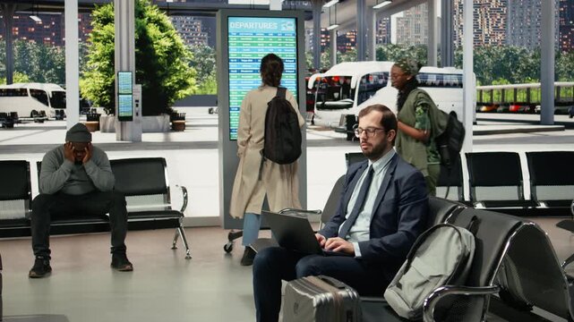 Frustrated business traveler works on a laptop in a bus station during delays, completing tasks on a notebook while slow public transit and a late coach extend the wait for departure and travel.