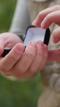 Beige outerwear fabric with stitching details visible. Left hand grips bottom edge of dark rectangular object. Stationary shot focuses on interaction with small item