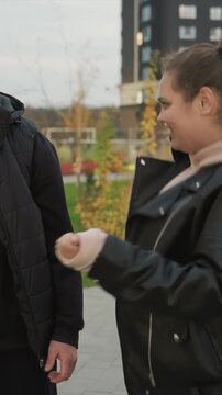 Woman in black leather jacket and man stand close together. Woman reaches out with open palm to man wearing puffer. Outdoor area features tall buildings and grassy ground