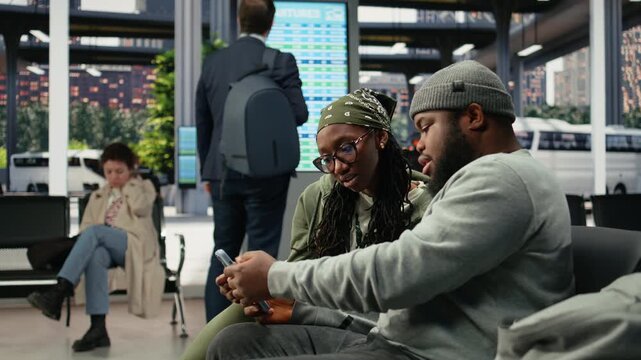 Happy travel companions use a smartphone in a coach station while catching up, checking their vacation itinerary together before departure.