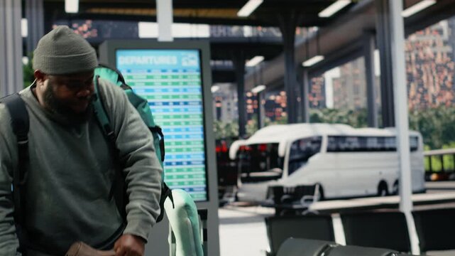 Tired passenger struggles with heavy baggage in a bus terminal before an intercity route, walking with bulky luggage trolleys toward a nearby bench.