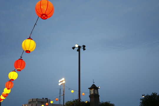 夕刻の曇り空に灯る祭り提灯。赤黄の装飾ランプと街灯。夜間イベント