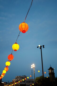 夕刻の曇り空に灯る祭り提灯。赤黄の装飾ランプと街灯。夜間イベント