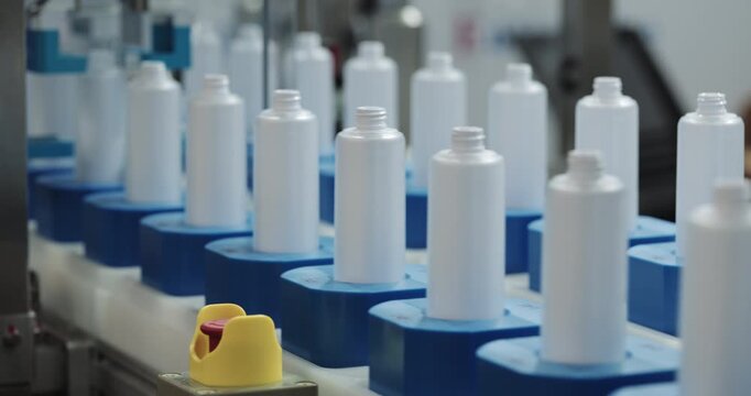 Automated equipment twists caps on blank opaque plastic bottles moving by conveyor belt. Production line packs liquid chemicals into containers at modern plant