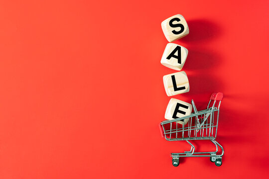 Miniature shopping cart filled with wooden letter blocks spelling SALE on a vibrant red background, symbolizing retail promotions, discounts, shopping, bargains, and seasonal sales.