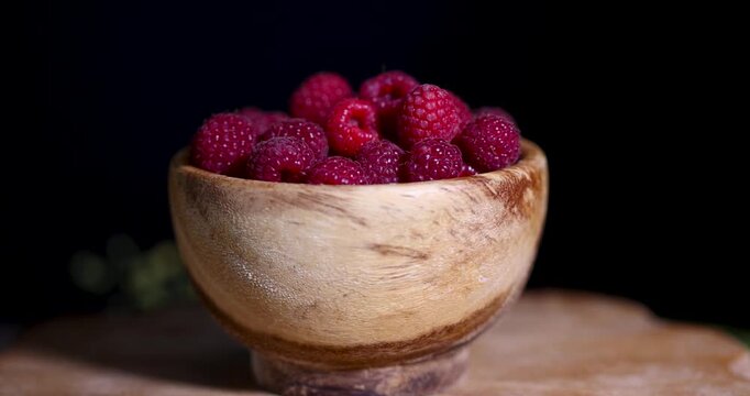 Fresh red raspberries in a bowl made of textured wood, a bowl of raspberries is placed on a black kitchen table next to the leaves.