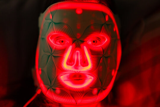 Woman wearing glowing red LED light therapy mask with geometric patterns, illustrating modern skincare technology and futuristic beauty treatment under vivid illumination.