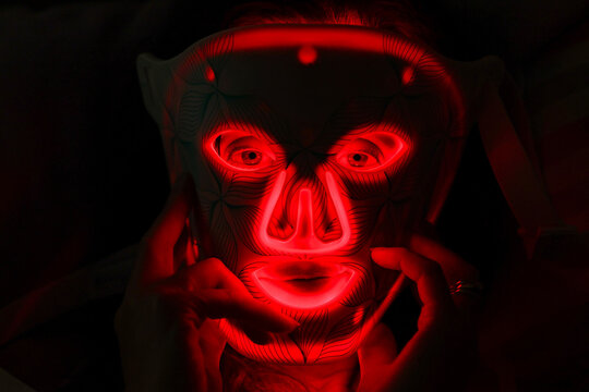 A person wears a glowing red LED light therapy mask with geometric patterns, illustrating modern skincare technology and futuristic beauty treatment under vivid illumination.
