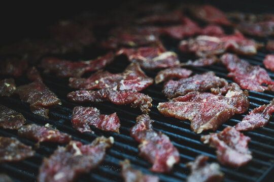Thin slices of round steak cook slowly on a pellet grill smoker, releasing aromatic smoke as they transform into tender homemade beef jerky under warm afternoon light.