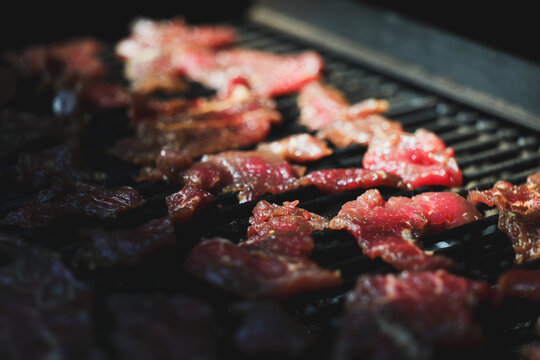 Thin slices of round steak cook slowly on a pellet grill smoker, releasing aromatic smoke as they transform into tender homemade beef jerky under warm afternoon light.