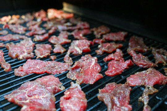 Slices of round steak rest on a pellet grill smoker, their glossy surface glistening in light as smoke rises, capturing the slow process of beef jerky preparation.