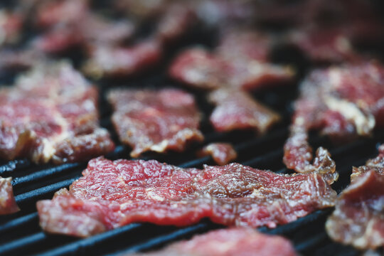 Thin slices of round steak cook slowly on a pellet grill smoker, releasing aromatic smoke as they transform into tender homemade beef jerky under warm afternoon light.