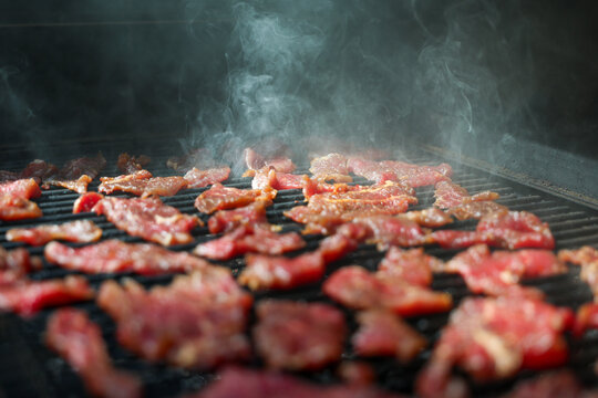 Thin strips of marinated round steak sizzle on a pellet grill smoker, surrounded by rising smoke and glowing heat, illustrating the art of homemade beef jerky.