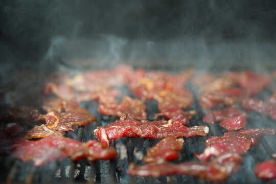 Thin strips of marinated round steak sizzle on a pellet grill smoker, surrounded by rising smoke and glowing heat, illustrating the art of homemade beef jerky.